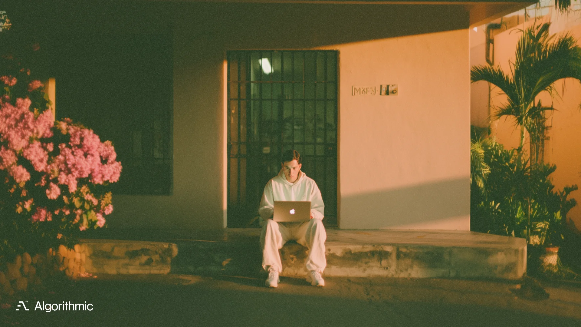 A young man in a white hoodie sitting on concrete steps outside a barred doorway, intensely focused on a laptop, bathed in warm golden light - representing a founder coding his way out of a trapped enterprise software position.